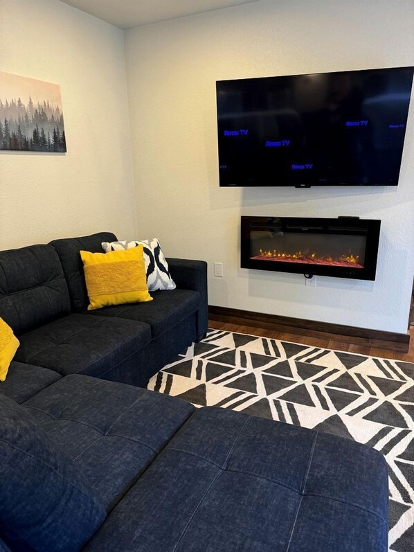 A living room with a dark-colored sofa, a flat-screen TV mounted above a fireplace, and a patterned rug on the floor.