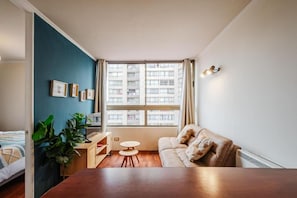 Ein modernes Wohnzimmer mit einer Couch, einem kleinen runden Tisch, einem Bett und einem großen Fenster mit Vorhängen.