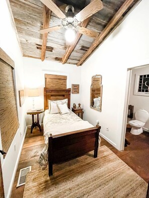 A bedroom with a wooden bed, a ceiling fan, a nightstand, a mirror, and a bathroom with a toilet.