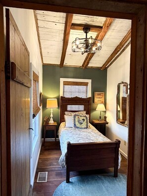 Un couloir étroit mène à une chambre à coucher où se trouvent un lit en bois, une table de chevet avec une lampe, un mur orné d’un miroir et un ventilateur de plafond.