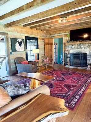 A rustic interior with a stone fireplace, wooden beams, a patterned rug, and a seating area with a wooden coffee table.