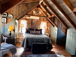 A rustic attic bedroom with a sloped ceiling, a bed with a quilted bedspread, a bedside table with a lamp, and a small chair.