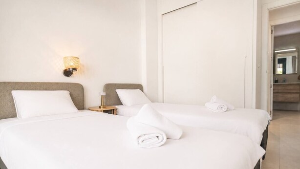 Habitación de hotel con dos camas, una mesita de noche con lámpara, y un baño visible a través de una puerta abierta.