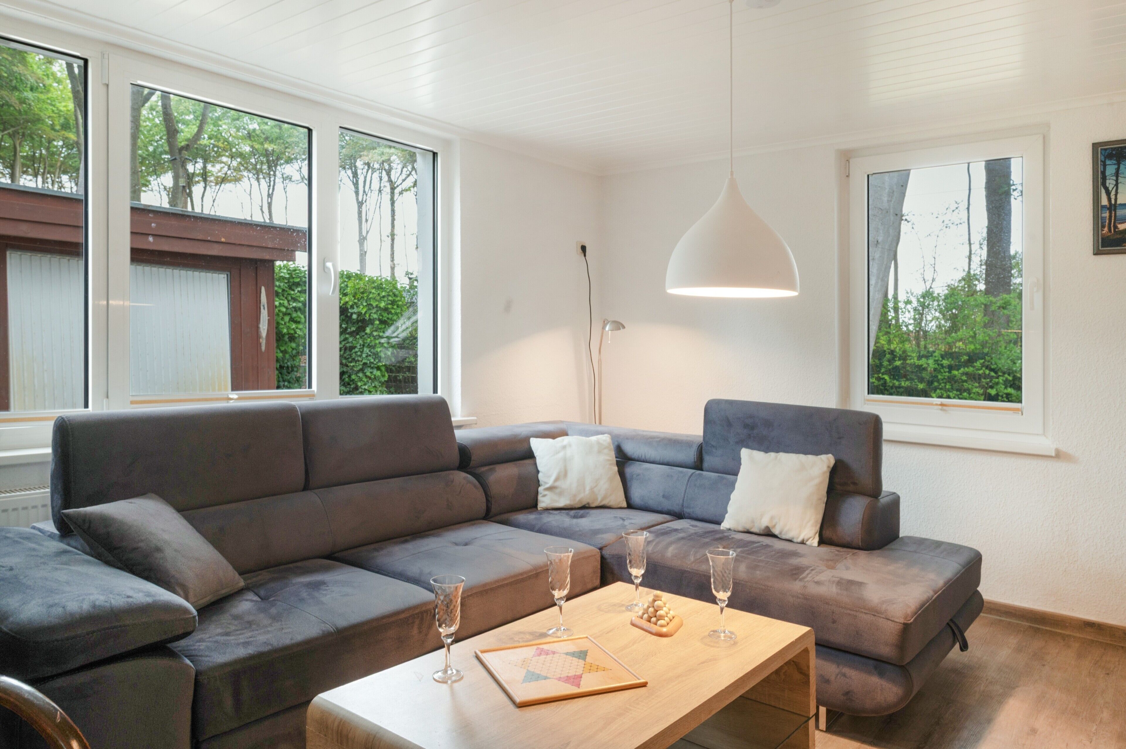 A modern living room with a sectional sofa, a coffee table, and two large windows offering a view of greenery.
