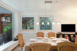 A dining area with a table set for four, two windows with views of greenery, a flat-screen TV, and a bookshelf with books. - Holiday Home for 6 Guests in Graal-Müritz (Baltic Sea Spa) (Graal-Müritz)