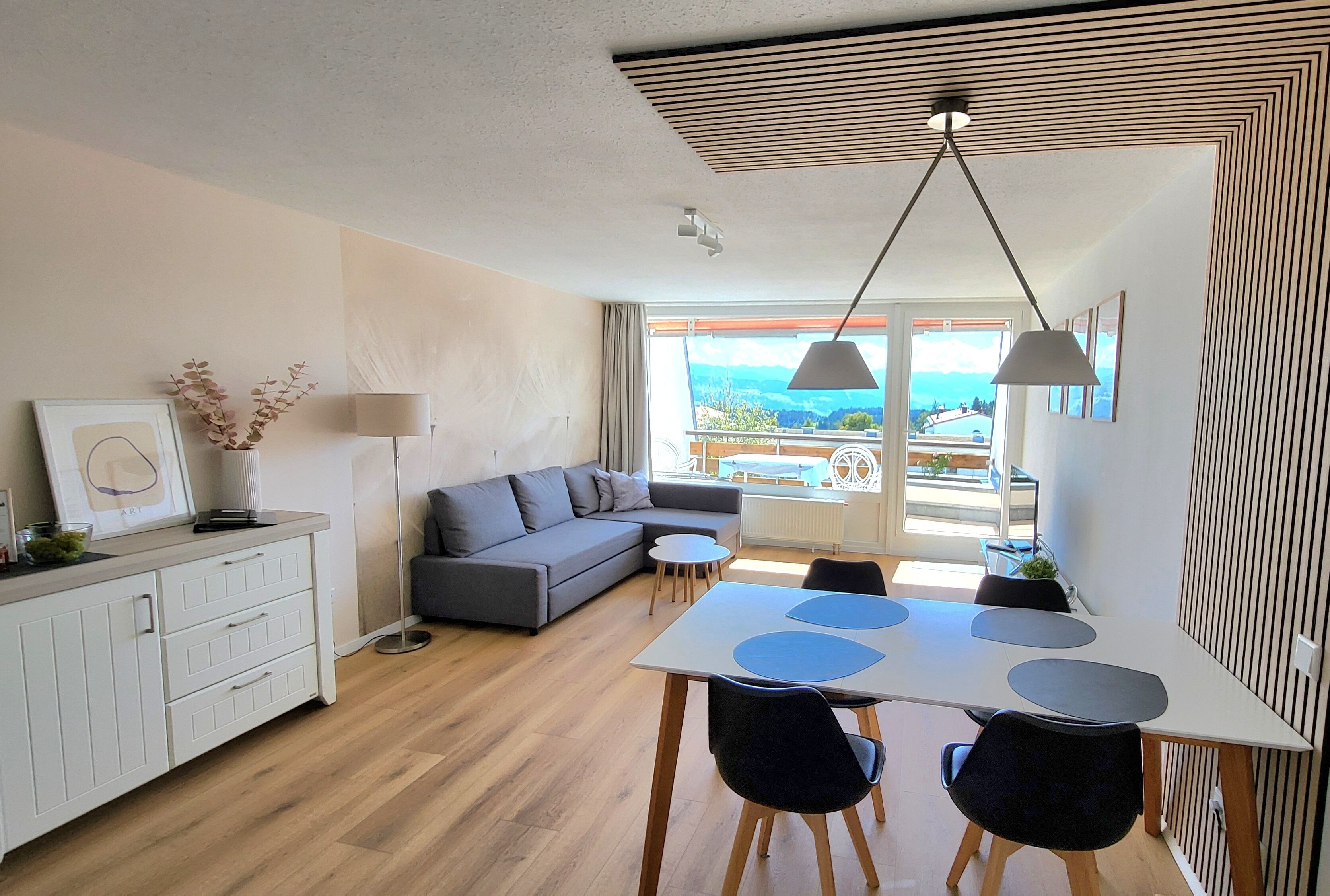 A modern living room with a grey sofa, a dining table with chairs, and a view of the outdoors through large windows.