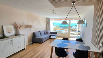 A modern living room with a grey sofa, a dining table with chairs, and a view of the outdoors through large windows.