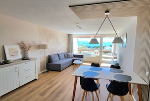 Ein modernes Wohnzimmer mit einem grauen Sofa, einem Esstisch mit Stühlen und Blick ins Freie durch große Fenster.