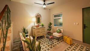 Une pièce avec un sol à carreaux, un ventilateur de plafond, une commode, une chaise avec un coussin à motifs de pastèque, une plante en pot et une valise.