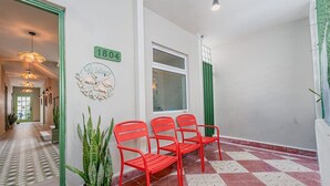 Um corredor com piso quadriculado, porta verde, placa com número e cadeiras vermelhas.