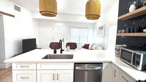 A modern kitchen with white cabinetry, a stainless steel dishwasher, a microwave, and a stove. There are two pendant lights, a sink, and a window with blinds. - Coronado Historic Casita · King Bed · Private Yard · Phoenix (Phoenix)