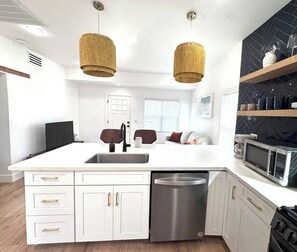 A modern kitchen with white cabinetry, a stainless steel dishwasher, a microwave, and a stove. There are two pendant lights, a sink, and a window with blinds. - Coronado Historic Casita · King Bed · Private Yard · Phoenix (Phoenix)