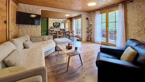En hyggelig stue med træloft, stenvægge, et fladskærms-tv og et spiseområde, der kan ses gennem en åben dør.