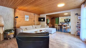 En hyggelig stue med træloft, sofa, en læderstol i sort farve, et fjernsyn og et køkken i baggrunden.