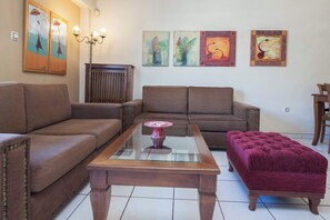A living room with a brown sofa, a glass-top coffee table, a red ottoman, and wall art. - Elegant 2 Bd Apt with Balcony in Korydallos Square (Koridallos)