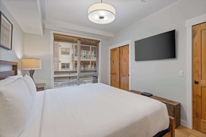 Ein Schlafzimmer mit einem Bett, einem an der Wand montierten Fernseher, Holztüren und einem Fenster mit Jalousien.