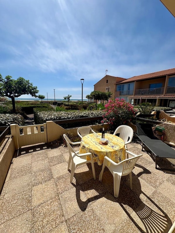 Eine Terrasse im Freien mit einem runden Tisch und Stühlen, Blick auf das Meer und ein Gebäude mit einem Balkon.
