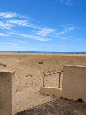 Ein sandiger Strand mit blauem Himmel, einem Metallgeländer und einigen Personen in der Ferne.