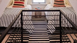 Un dormitorio acogedor en el ático, con techo inclinado, una ventana con persianas, una cama con ropa de cama a rayas y una lámpara colgante.