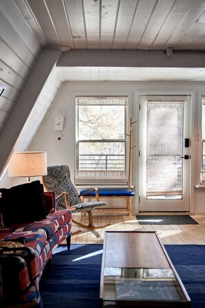 A room with a sloped ceiling, a sofa, a chair, a table, and two windows with blinds.