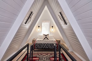 A triangular attic bedroom with a bed, a window, and two wall-mounted light fixtures.
