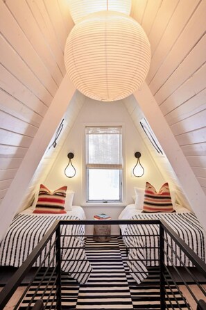 A cozy attic bedroom with a sloped ceiling, a window with blinds, a bed with striped bedding, and a hanging light fixture.