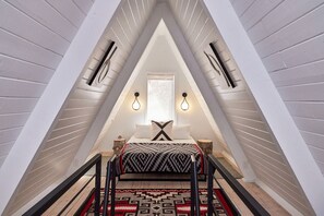 A triangular attic bedroom with a bed, a window, and two wall-mounted light fixtures. - Cool A-Frame Cabin Rental in New Mexico, for Unforgettable Vacations on the Pecos River (Pecos)