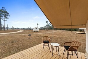 Una terraza de madera con dos sillas de mimbre y una mesita, un edificio blanco al fondo y un cielo despejado.
