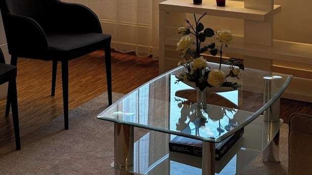 Una habitación con un ventanal grande, una silla negra, una mesa de vidrio con un florero y un estante blanco.