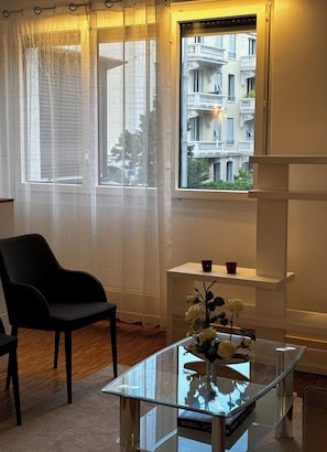 Ein Zimmer mit großem Fenster, einem schwarzen Stuhl, einem Glastisch mit einer Vase und einem weißen Regal.