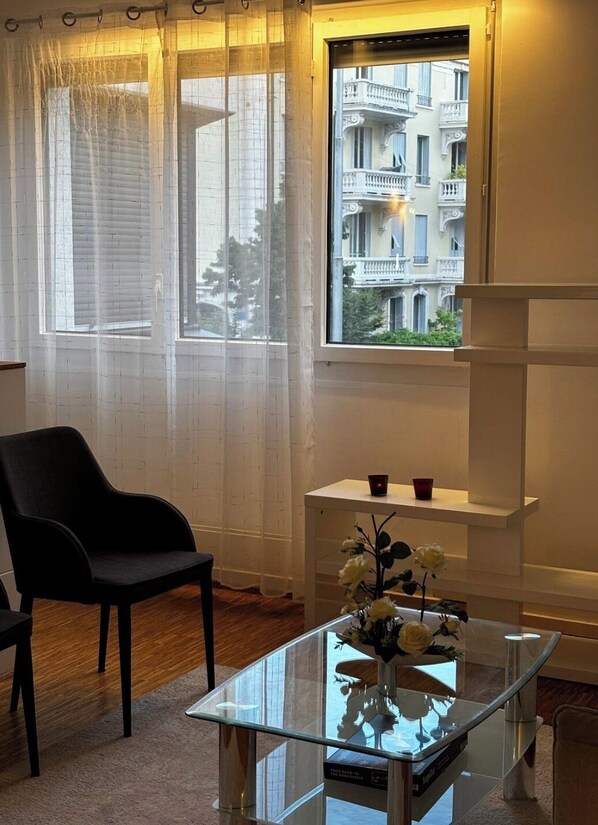 A room with a large window, a black chair, a glass table with a vase, and a white shelf.