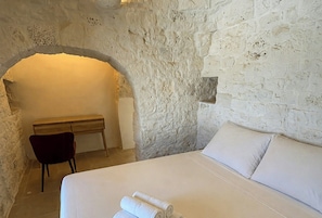A bedroom with a stone wall, a bed with white linens, a desk, and a chair. - Trullo with infinity pool amid olive groves (Alberobello)