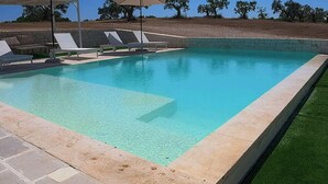 A swimming pool with clear blue water, surrounded by lounge chairs and umbrellas, and a stone wall in the background. - Trullo with infinity pool amid olive groves (Alberobello)