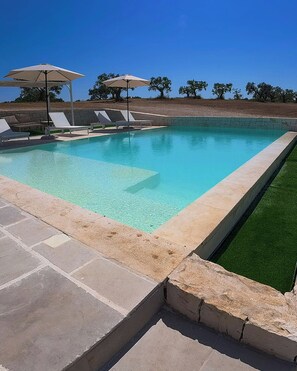 Ein Swimmingpool mit klarem, blauem Wasser, umgeben von Liegestühlen und Sonnenschirmen, mit einer Steinmauer im Hintergrund.