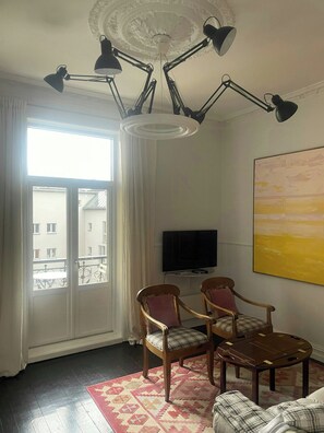 A living room with a large window, a ceiling light fixture, a television, and a painting on the wall. - Classic flat with balcony in St. Hanshaugen (Oslo)