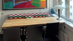 Ein Zimmer mit einem großen, farbenfrohen Gemälde, einem Holztisch mit einer karierten Bank, einer Lampe und einem Fenster mit Blick auf Gebäude.
