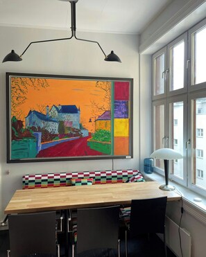 Una stanza con un quadro colorato di grandi dimensioni, un tavolo in legno con una panca a scacchi, una lampada e una finestra che si affaccia su degli edifici.