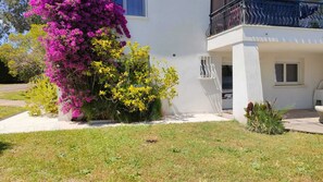 Ein weißes Gebäude mit Balkon, ein Garten mit rosa und gelben Blumen und ein klarer Himmel.