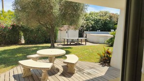 Eine Terrasse mit rundem Tisch und Bänken, umgeben von Grünpflanzen und einem weißen Gebäude im Hintergrund.