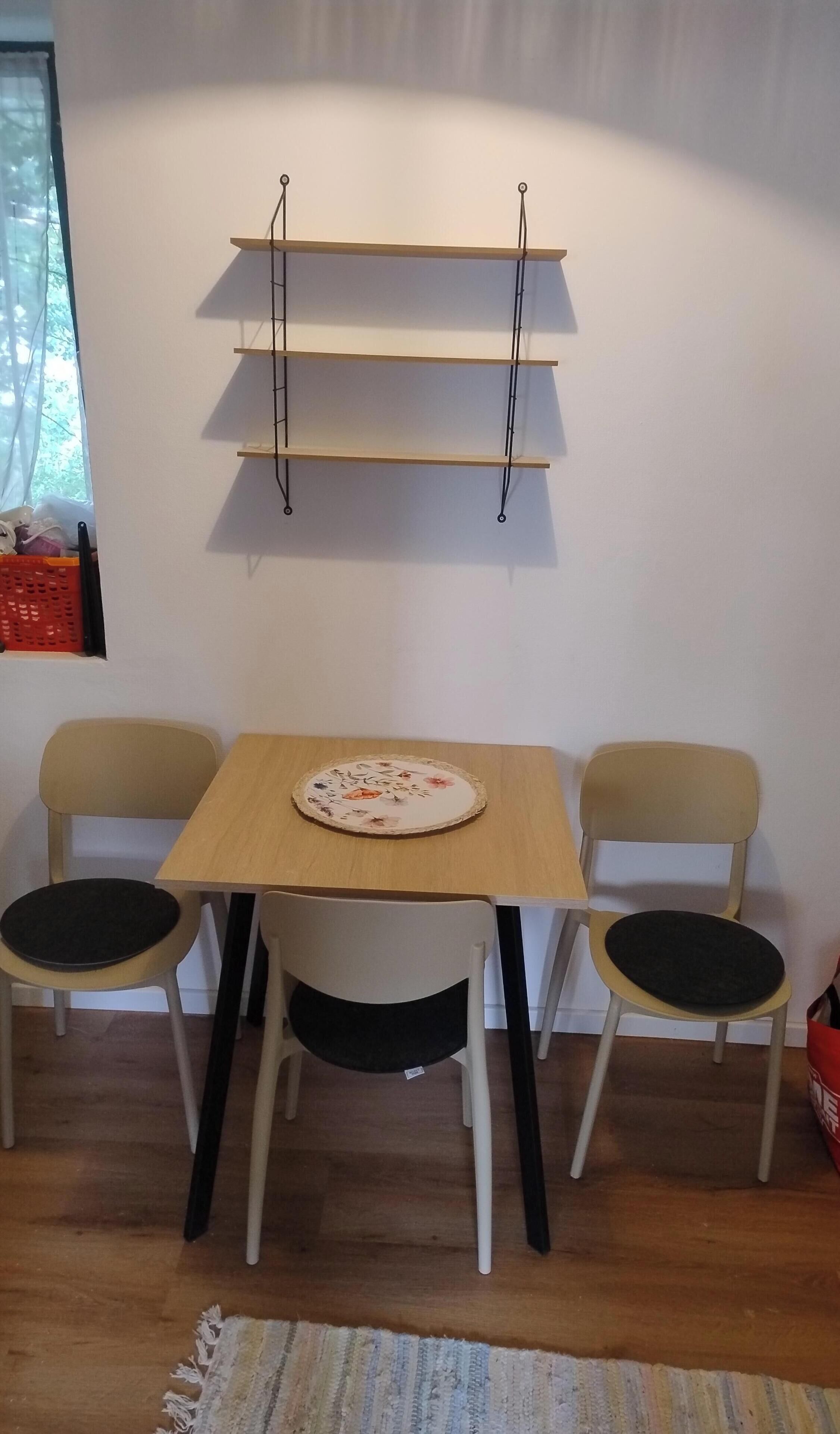 Ein kleiner Essbereich mit einem Holztisch und zwei Stühlen, einem darüber angebrachten Regal und einem Fenster mit Vorhängen.