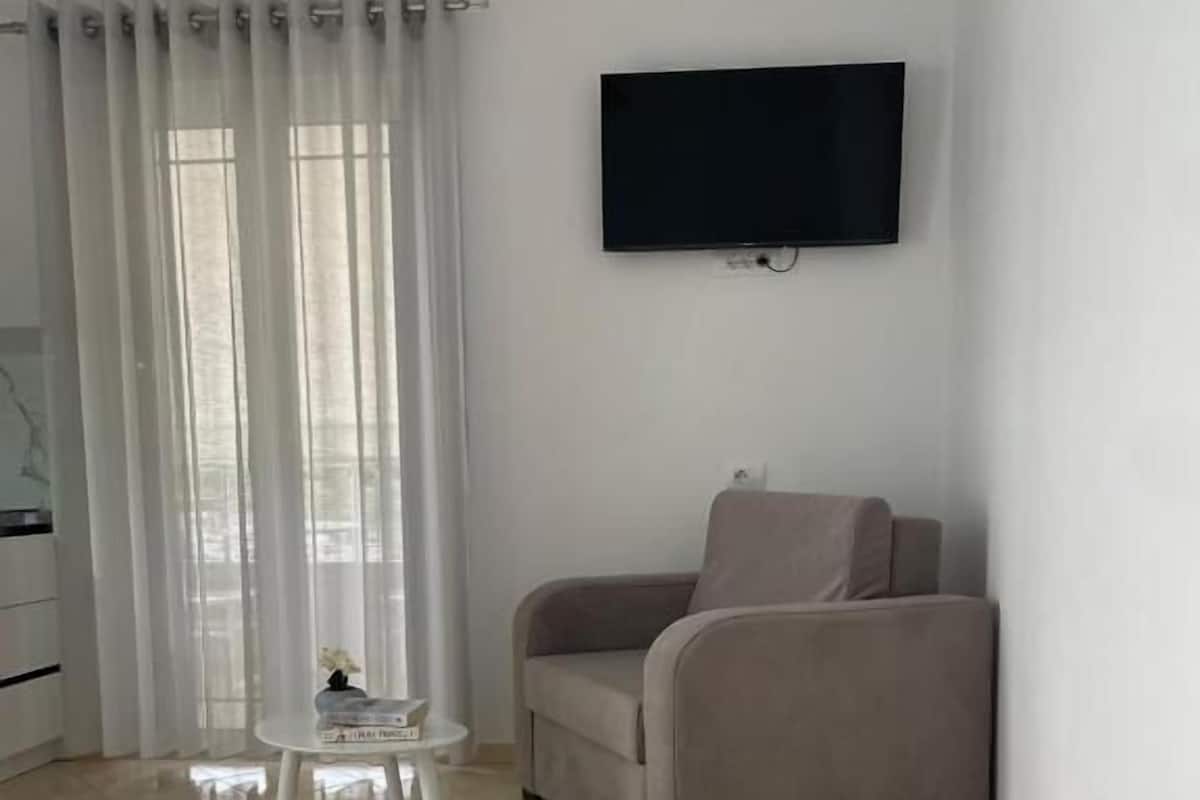 A hotel room with a bed, a TV mounted on the wall, a chair, and a small table with a book and a rose.