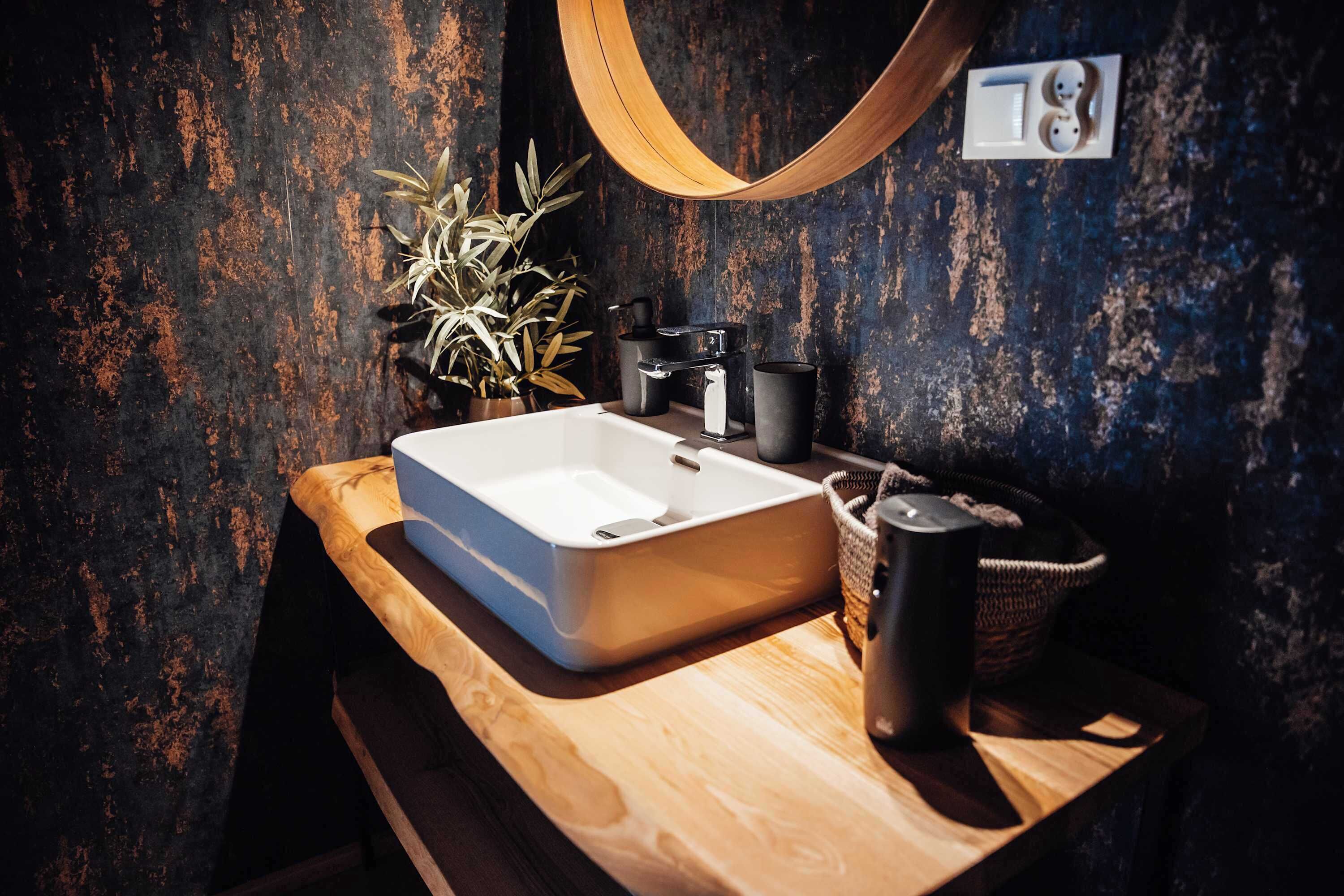 Ein Badezimmer mit einem Holzkosmetikschrank, einem weißen Waschbecken, einem schwarzen Wasserhahn, einer Topfpflanze, einem gewebten Korb und einem Handtuchhalter.