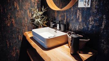 Ein Badezimmer mit einem Holzkosmetikschrank, einem weißen Waschbecken, einem schwarzen Wasserhahn, einer Topfpflanze, einem gewebten Korb und einem Handtuchhalter.
