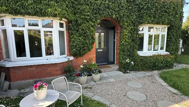 Una casa con pareti ricoperte d'edera, facciata in mattoni e una porta d'ingresso. Un piccolo giardino con tavolo e sedie, e un vialetto che conduce alla porta.