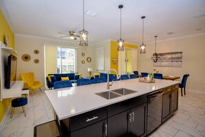 A modern kitchen with a central island, black cabinets, and a white countertop. There are blue chairs and a yellow sofa in the adjacent living area. - Go!#28 Champions Gate (Kissimmee)
