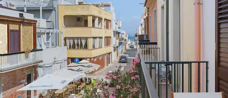A narrow street with buildings on both sides, outdoor seating with umbrellas, and a view of the sea in the distance.