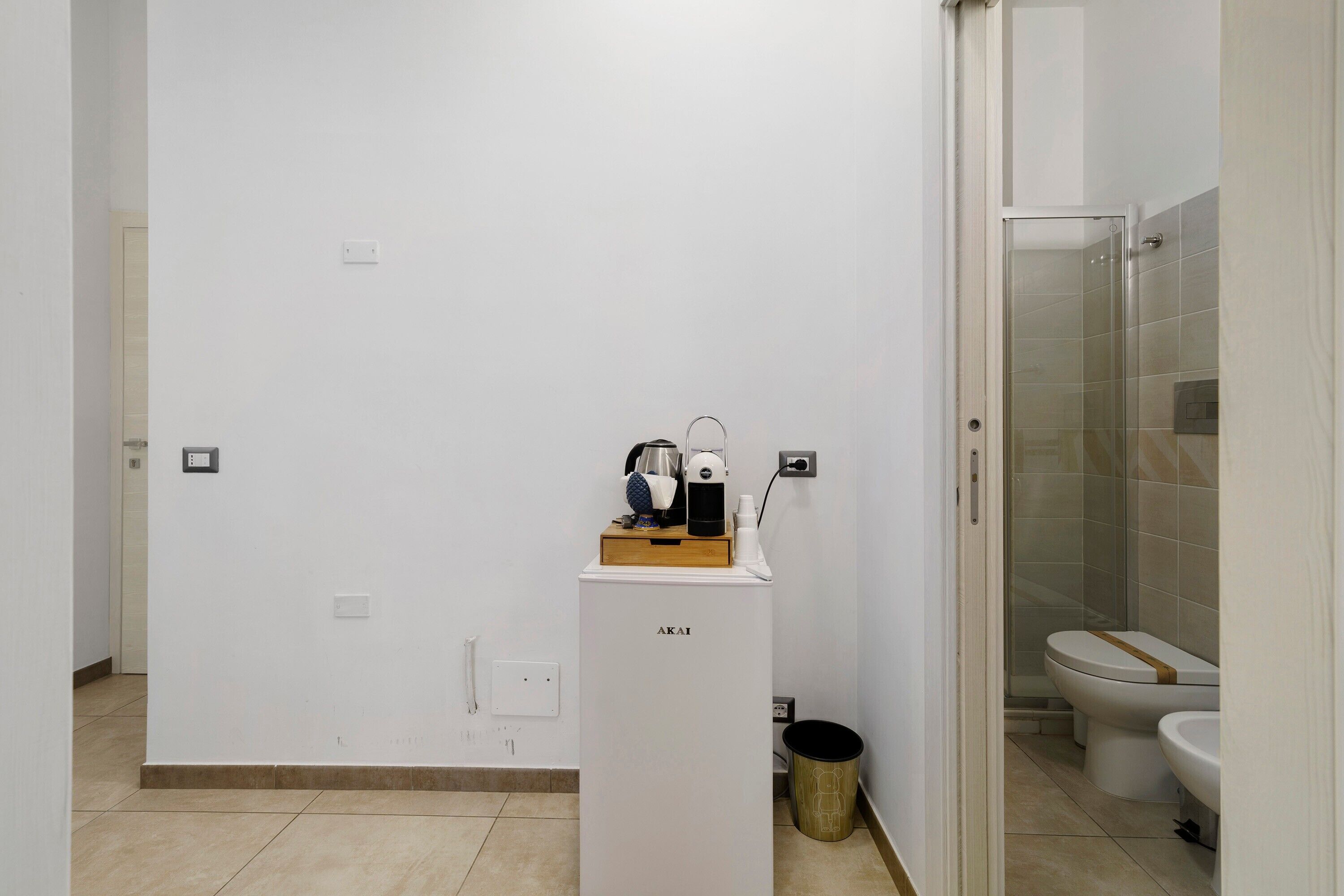 Baño pequeño y limpio con inodoro, ducha y lavamanos. Hay un pequeño cubo de basura y una máquina de café en un estante.