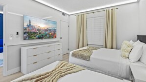 Hotellihuone, jossa on suuri litteänäyttöinen televisio, kaksi sänkyä, valkoinen lipasto ja ikkuna, jossa on verhot.