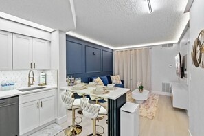 Eine moderne Küche mit weißen Schränken, ein Essbereich mit einem weißen Tisch und goldenen Stühlen sowie eine blaue Akzentwand.