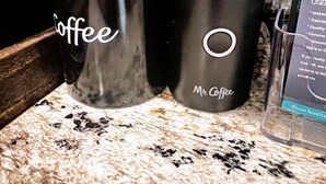 Zwei schwarze Trinkbecher mit dem Aufdruck 'Coffee' und der Marke 'Mr. Coffee' auf einer Küchenarbeitsplatte.